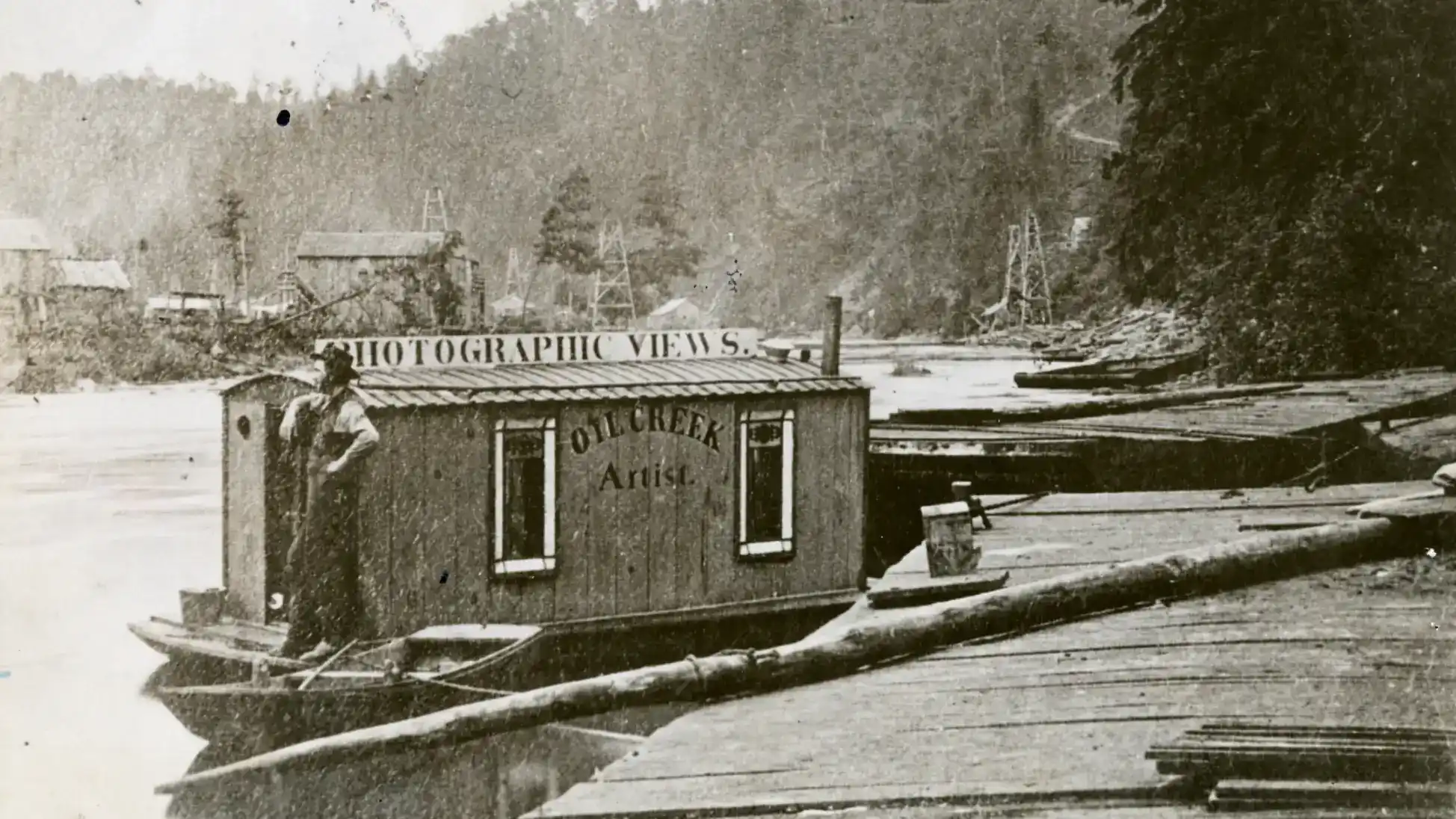 John Mather’s floating photography studio on Oil Creek, Pennsylvania, in the early 1860s, labeled “Photographic Views” and “Oil Creek Artist,” with oil derricks nearby.
