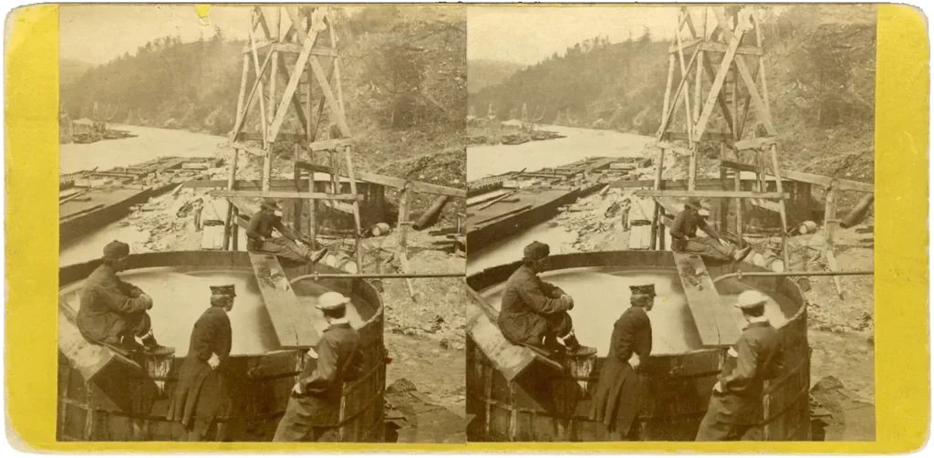 Historic stereograph image showing a flowing oil well at Oil Creek, Pennsylvania, with several men observing oil-filled wooden barrels and early drilling equipment.